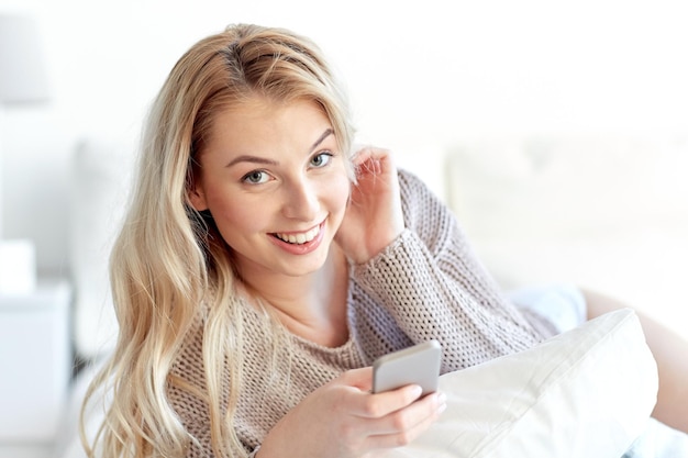 concept de technologie, de communication et de personnes - heureuse jeune femme allongée dans son lit avec un smartphone dans la chambre à coucher