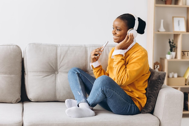 Concept de technologie et de loisirs des gens heureuse jeune femme afro-américaine assise sur un canapé avec smartphone et casque écoutant de la musique à la maison