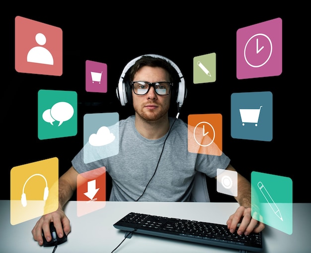 concept de technologie, de médias et de personnes - jeune homme ou pirate dans un casque et des lunettes assis à table avec un clavier d'ordinateur pc et des icônes de menu sur un écran virtuel