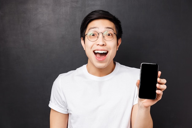 Concept de technologie, de messagerie et de personnes. Portrait de gros plan de jeune homme asiatique heureux, surpris et impressionné en t-shirt blanc, montrant l'écran du smartphone étonné avec une application cool