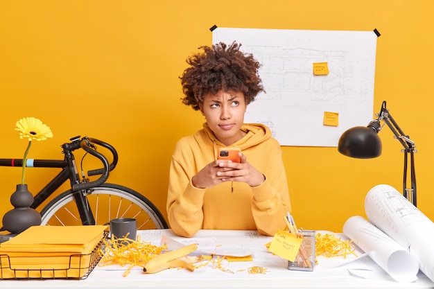 Photo concept de technologie d'occupation de personnes. un étudiant afro-américain réfléchi tient un smartphone avec un look frustré vêtu d'un sweat-shirt travaille sur un projet architectural de la maison a un désordre sur le bureau