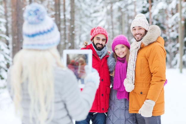 concept de technologie, de saison, d'amitié et de personnes - groupe d'hommes et de femmes souriants prenant des photos avec un ordinateur tablette dans la forêt d'hiver