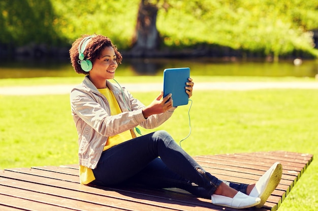 concept de technologie, de style de vie et de personnes - jeune femme ou adolescente afro-américaine souriante avec ordinateur tablette et écouteurs écoutant de la musique ou regardant des vidéos dans le parc d'été