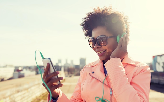 concept de technologie, de style de vie et de personnes - jeune femme ou adolescente afro-américaine souriante avec smartphone et écouteurs écoutant de la musique à l'extérieur