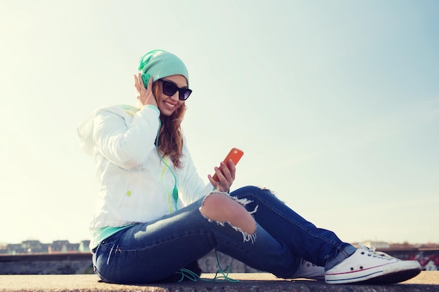 concept de technologie, de style de vie et de personnes - jeune femme ou adolescente souriante avec smartphone et écouteurs écoutant de la musique à l'extérieur