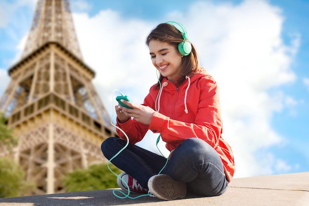 concept de technologie, de voyage, de tourisme et de personnes - jeune femme souriante ou adolescente avec smartphone et écouteurs écoutant de la musique sur fond de tour eiffel