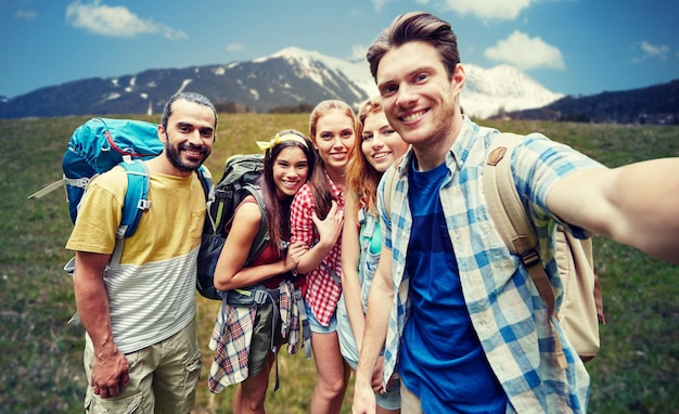 concept de technologie, de voyage, de tourisme, de randonnée et de personnes - groupe d'amis souriants marchant avec des sacs à dos prenant un selfie par smartphone ou appareil photo sur fond naturel