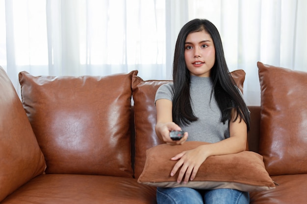 Concept de télévision et de bonheur. Belle femme asiatique décontractée assise sur un canapé dans le salon, tenant la télécommande de la télévision et regardant quelque chose avec un visage souriant heureux. Concept de mode de vie.