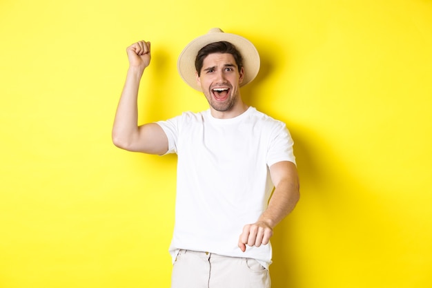 Concept de tourisme et d'été. Jeune homme voyageur montrant un geste de rodéo, debout dans un chapeau de paille et des vêtements blancs, debout sur fond jaune.