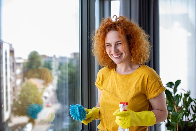 Concept de travaux ménagers et d'entretien ménager femme heureuse dans des gants nettoyant la fenêtre avec un chiffon et un spray nettoyant à la maison