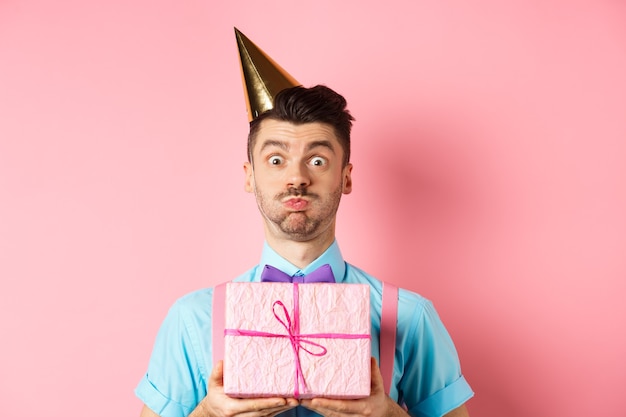 Concept De Vacances Et De Célébration. Drôle De Gars Regardant La Caméra Surpris, Portant Un Chapeau De Fête, Tenant Un Cadeau D'anniversaire Et Retenant Son Souffle, Faisant La Moue à La Caméra, Fond Rose.