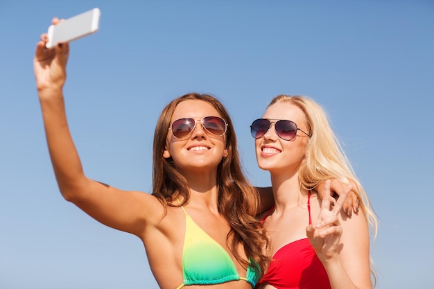 concept de vacances d'été, de vacances, de voyage, de technologie et de personnes - deux jeunes femmes souriantes sur la plage faisant du selfie avec un smartphone sur fond de ciel bleu