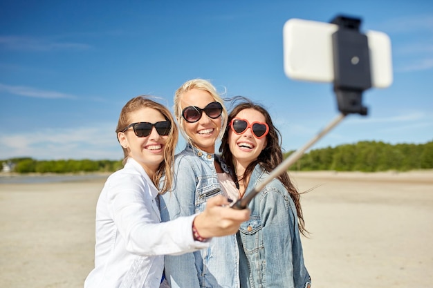 concept de vacances d'été, de vacances, de voyage, de technologie et de personnes - groupe de jeunes femmes souriantes prenant des photos avec un smartphone sur un bâton de selfie sur la plage