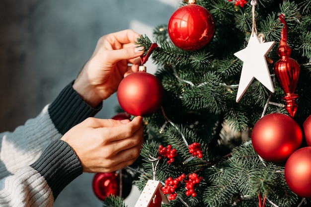 Concept de vacances d'hiver. Mains d'homme décorant le sapin vert avec des ornements de boule rouge.