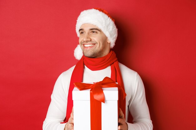 Concept de vacances d'hiver, Noël et mode de vie. Gros plan sur un mec séduisant en bonnet et écharpe, souriant et regardant à gauche tout en tenant le cadeau du nouvel an, debout sur fond rouge.