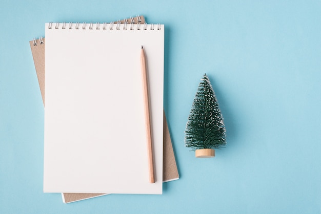 Concept de vacances de Noël. En haut au-dessus de la vue aérienne, photo à plat d'un cahier vierge et d'un petit arbre de Noël isolé sur fond de couleur bleu pastel avec fond