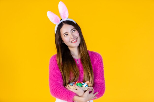 Concept de vacances de Pâques, heureuse jeune femme asiatique portant des oreilles de lapin main tenant un panier avec des oeufs de Pâques colorés en isolé sur fond de studio espace copie vierge jaune.