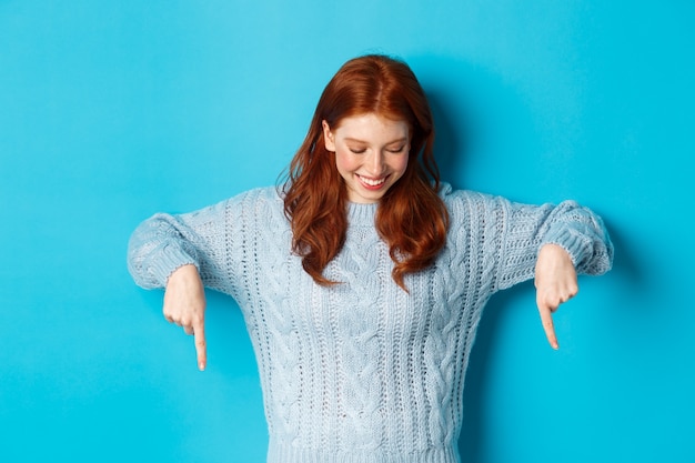 Concept de vacances et de personnes d'hiver. Joyeuse rousse en pull, pointant les doigts vers le bas et regardant heureux au logo, debout sur fond bleu