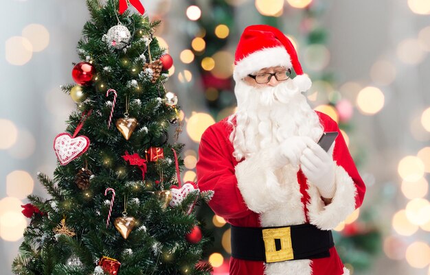 concept de vacances, de technologie et de personnes - homme en costume de père noël avec smartphone et arbre de noël sur fond de lumières bleues