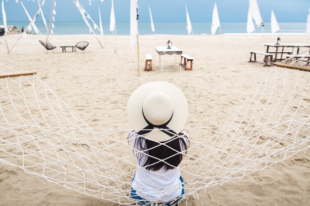 Concept de vacances de voyage d'été, heureux voyageur solo femme asiatique avec chemise sans manches blanche et chapeau se détendre dans un hamac sur la plage tropicale à Hua Hin, Thaïlande