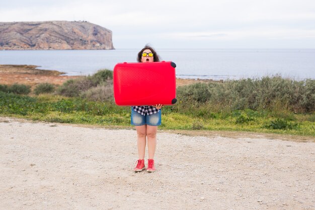 Concept de vacances, de voyage et de tourisme. jeune femme avec une valise rouge sur fond de mer.