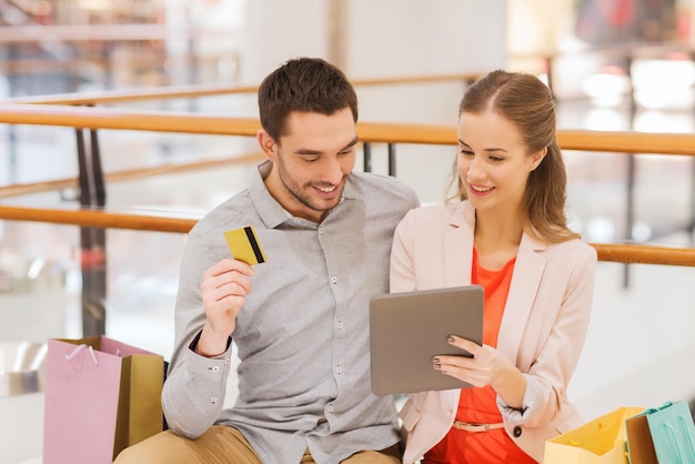 concept de vente, de consommation, de loisirs et de personnes - couple heureux avec ordinateur tablette, carte de crédit et sacs à provisions dans le centre commercial