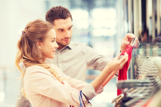 concept de vente, de consommation et de personnes - jeune couple heureux avec des sacs à provisions choisissant une robe dans un centre commercial