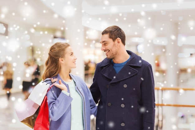 concept de vente, de consommation et de personnes - jeune couple heureux avec des sacs à provisions parlant dans un centre commercial avec effet de neige