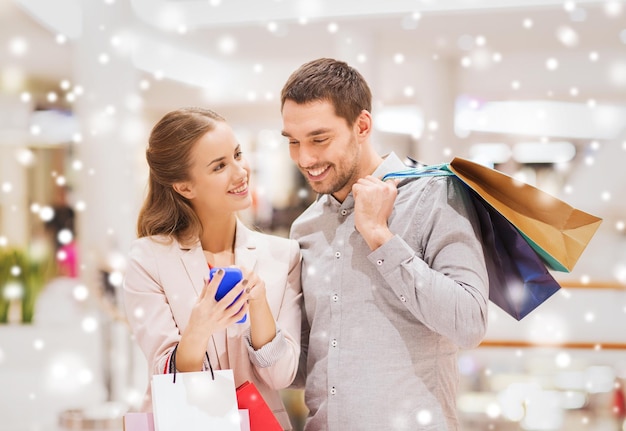 concept de vente, de consommation, de technologie et de personnes - jeune couple heureux avec des sacs à provisions et un smartphone parlant dans un centre commercial avec effet de neige