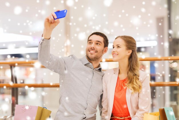 concept de vente, de consommation, de technologie et de personnes - jeune couple heureux avec des sacs à provisions et un smartphone prenant un selfie dans un centre commercial avec effet de neige