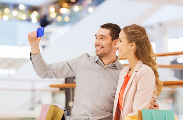 concept de vente, de consommation, de technologie et de personnes - jeune couple heureux avec des sacs à provisions et un smartphone prenant un selfie dans un centre commercial