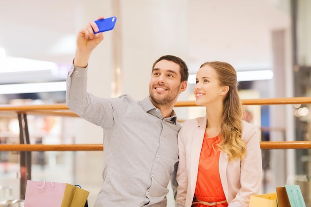 concept de vente, de consommation, de technologie et de personnes - jeune couple heureux avec des sacs à provisions et un smartphone prenant un selfie dans un centre commercial