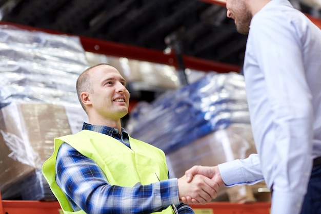 Photo concept de vente en gros, de logistique, de personnes et d'exportation - travailleur manuel et hommes d'affaires avec presse-papiers se serrant la main et faisant affaire à l'entrepôt