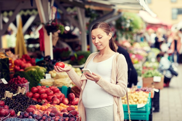 concept de vente, de shopping, de nourriture, de grossesse et de personnes - femme enceinte heureuse avec smartphone choisissant des légumes au marché de rue