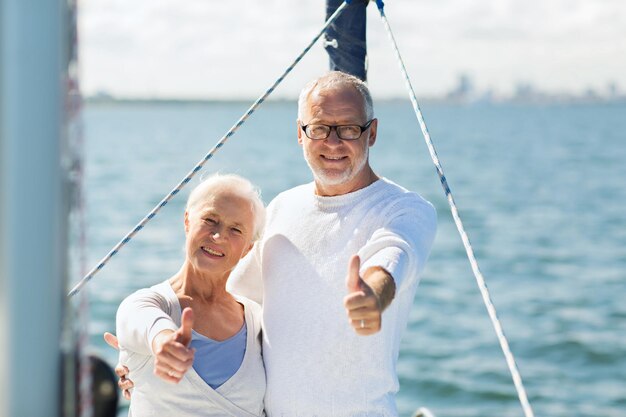 concept de voile, d'âge, de tourisme, de voyage et de personnes - heureux couple de personnes âgées embrassant sur un voilier ou un pont de yacht flottant et montrant les pouces vers le haut en mer