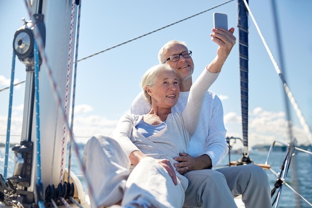 concept de voile, de technologie, de tourisme, de voyage et de personnes - couple de personnes âgées heureux avec un smartphone prenant un selfie sur un voilier ou un pont de yacht flottant en mer