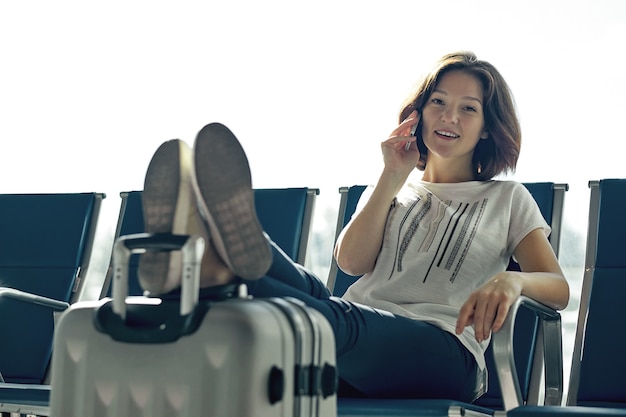 Concept de voyage aérien avec une jeune fille décontractée assise avec une valise à bagages à main. Femme de l'aéroport parlant au téléphone à la porte en attente dans le terminal.