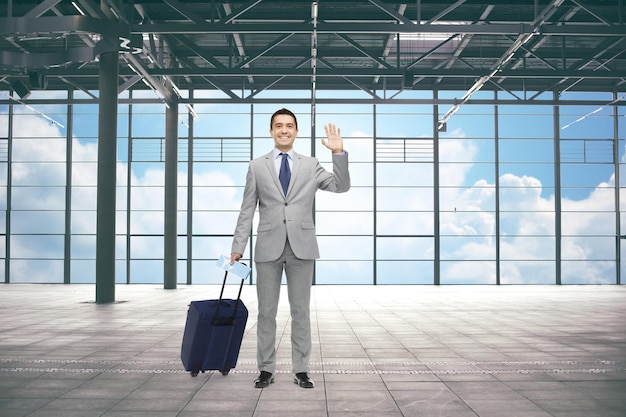 concept de voyage d'affaires, de voyage, de geste et de personnes - homme d'affaires heureux en costume avec sac de voyage agitant la main sur l'arrière-plan du terminal de l'aéroport