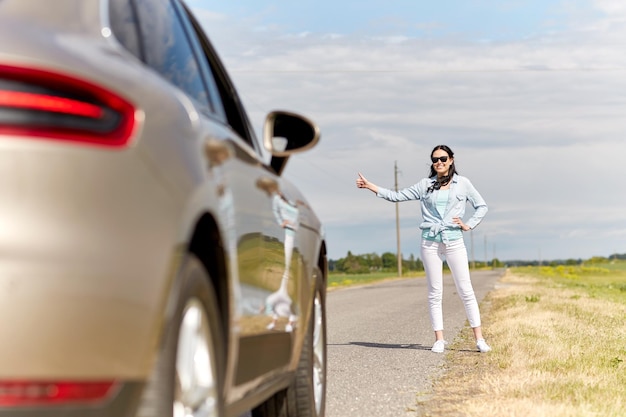 Concept de voyage, d'auto-stop, de voyage, de geste et de personnes - femme faisant de l'auto-stop et arrêtant une voiture avec un geste de pouce levé sur une route de campagne