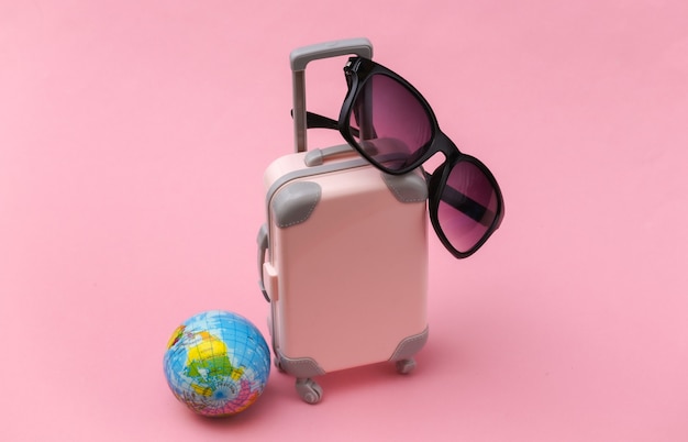 Concept de voyage international. Mini valise de voyage en plastique, globe et lunettes de soleil sur fond rose. Style minimaliste