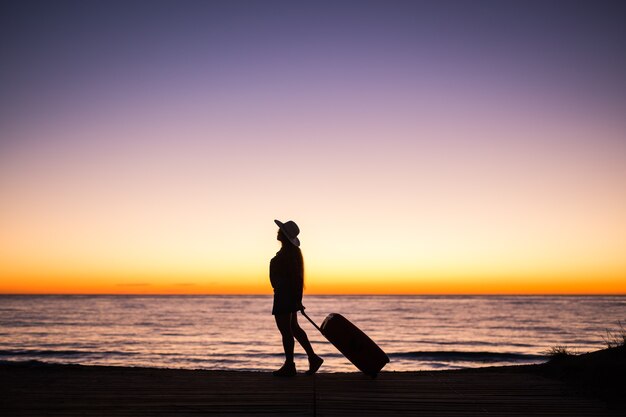 Concept de voyage, de plage et de vacances - silhouette féminine marchant le long de la côte de l'océan tirant la valise.