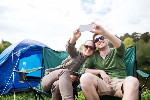 concept de voyage, de randonnée, de technologie, de tourisme et de personnes - couple souriant de voyageurs prenant un selfie par smartphone au camping