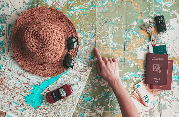 Photo concept de voyage sur la route à plat. main pointant vers un endroit sur la carte. chapeau, lunettes de soleil, petite voiture, passeport, argent et clés sur la carte routière. planification des vacances.