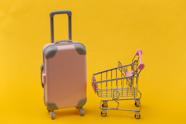 Concept de voyage et de shopping. Mini valise de voyage en plastique rose et chariot de supermarché sur fond jaune. Style minimaliste