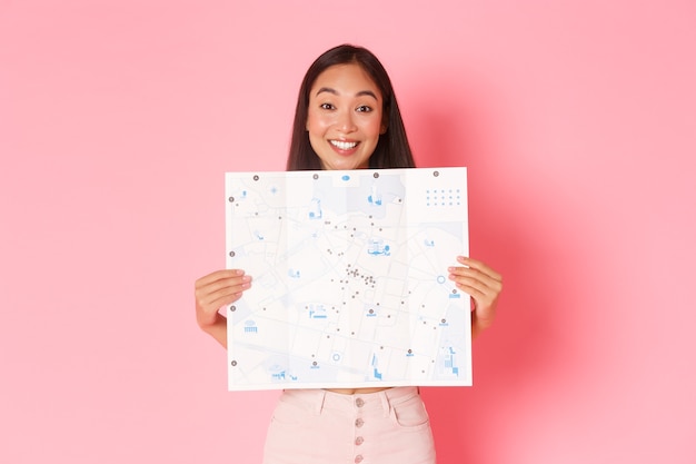 Concept de voyage, de style de vie et de tourisme. Enthousiaste et attrayante, une fille asiatique touriste explore la nouvelle ville, visite des musées, montre la carte de la ville avec des visites touristiques et souriant optimiste, mur rose