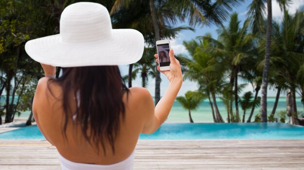 concept de voyage, de tourisme, d'été, de technologie et de personnes - souriante jeune femme ou adolescente en chapeau de soleil prenant selfie avec smartphone sur la plage de la station balnéaire avec palmiers et fond de piscine