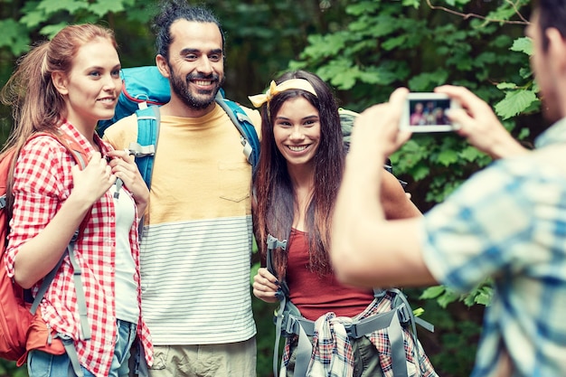 concept de voyage, de tourisme, de randonnée, de technologie et de personnes - groupe d'amis souriants marchant avec des sacs à dos prenant des photos par smartphone dans les bois