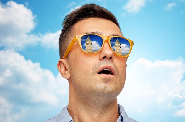 concept de voyage, de tourisme, de tourisme, d'émotions et de personnes - visage d'homme à lunettes de soleil regardant la tour big ben sur fond bleu ciel et nuages