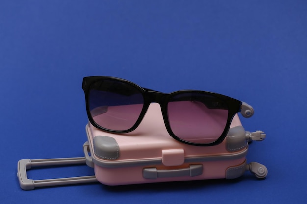 Concept de voyage ou de voyage. Mini valise de voyage en plastique avec lunettes de soleil sur fond bleu classique. Couleur 2020