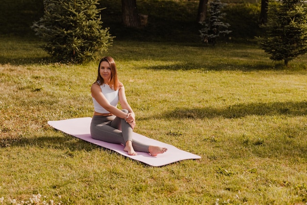 Concept de yoga pleine longueur femme mince sportive en t-shirt blanc et leggins gris assis avec le dos droit et les jambes croisées sur un tapis de pilates à l'extérieur au fond du parc en soirée